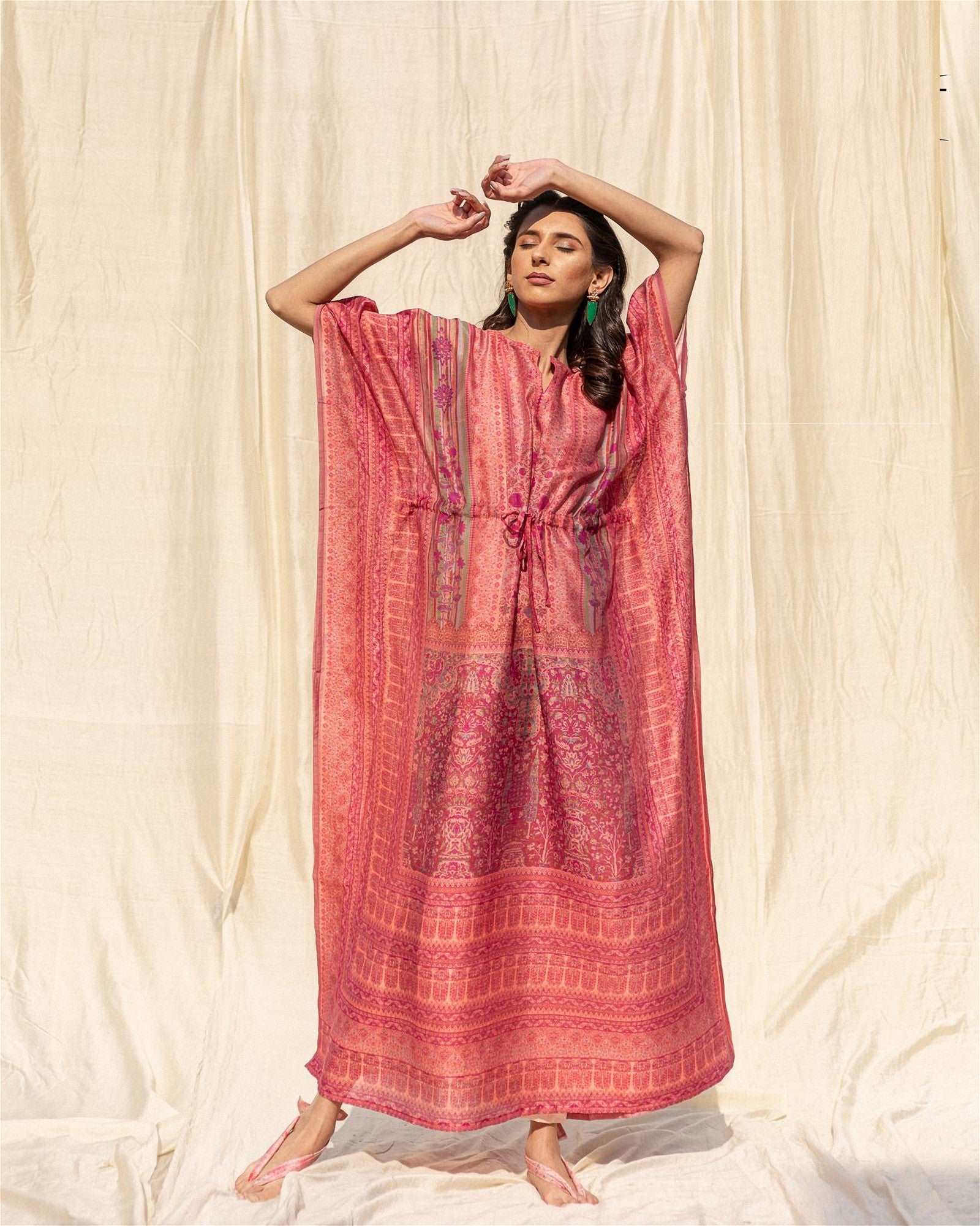 The model in the image is wearing Pink Soft Silk Premium Kaftan from Alice Milan. Crafted with the finest materials and impeccable attention to detail, the Kaftan / Dress looks premium, trendy, luxurious and offers unparalleled comfort. It’s a perfect clothing option for loungewear, resort wear, party wear or for an airport look. The woman in the image looks happy, and confident with her style statement putting a happy smile on her face.